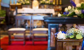 A floral bouquet and ribbons adorn a wooden pew in the foreground; behind, a red carpet leads to an altar in a decorated church, featuring chairs and various ornate elements.