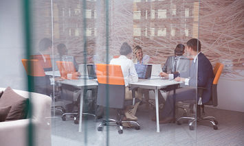 People are seated at a conference table, discussing and using laptops, inside a glass-walled office with modern decor, including orange chairs and wall art.