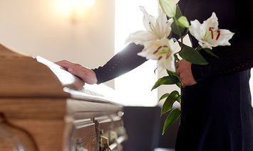 A person in black clothing holds white lilies, resting hand on a closed wooden casket in a softly lit room, implying a solemn or memorial setting.