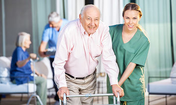 An elderly man using a walker is assisted by a smiling female healthcare worker in green scrubs; they are outdoors, with another elderly couple and healthcare worker in the background.