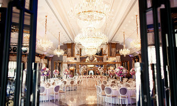 Opulent banquet hall adorned with grand chandeliers, elegantly set tables with floral arrangements, and high ceilings. The setting is luxurious, with ornate decorations and large windows